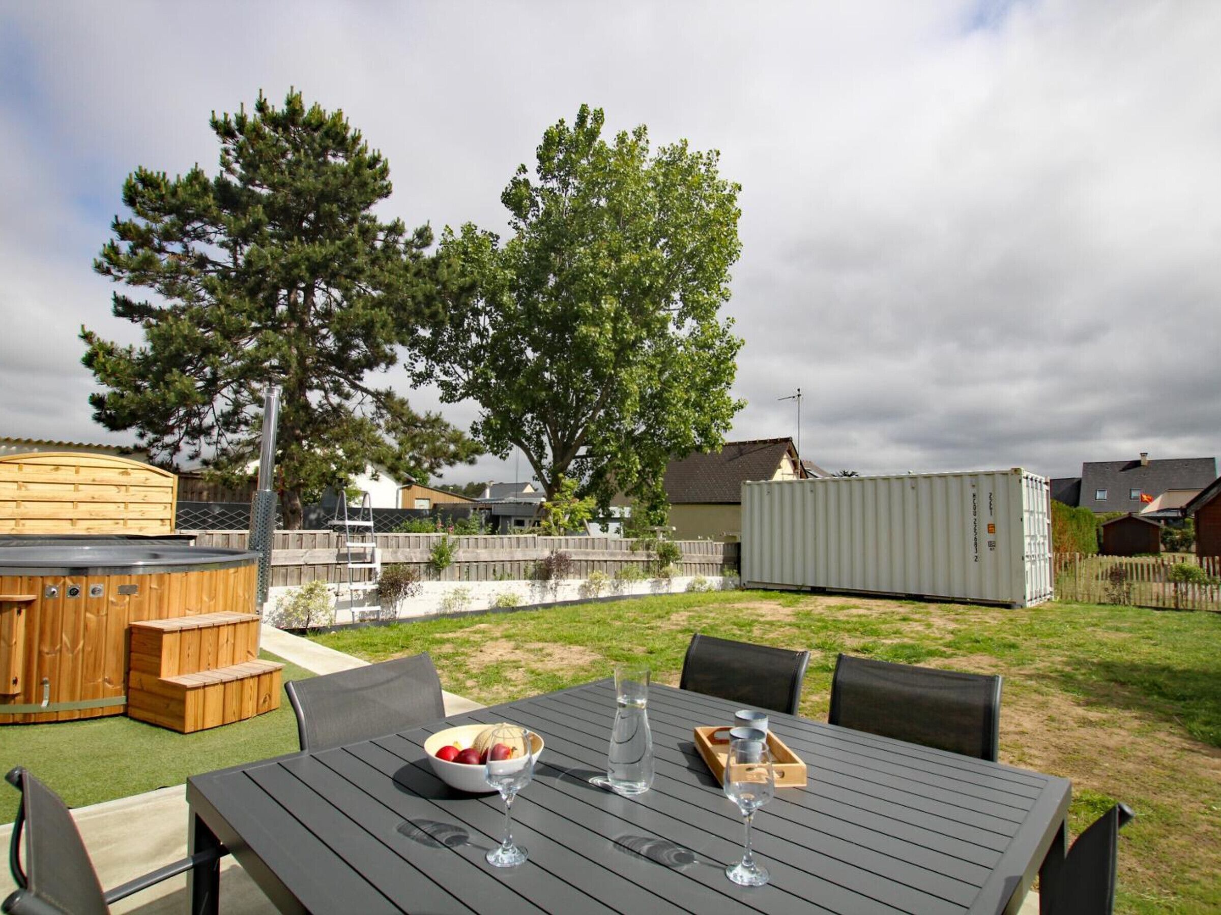 An outdoor patio with a table set for dining, surrounded by chairs, a wooden deck, and a large tree.