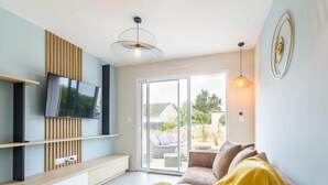 A modern living room with a flat-screen TV mounted on a wooden panel, a sofa with a yellow throw, and a sliding glass door leading to an outdoor patio. - Charming Modern Bungalow with Nordic Hot Tub, Near Beach and Amenities in Saint-Jean-de-la-Rivière (Saint-Jean-de-la-Rivière)