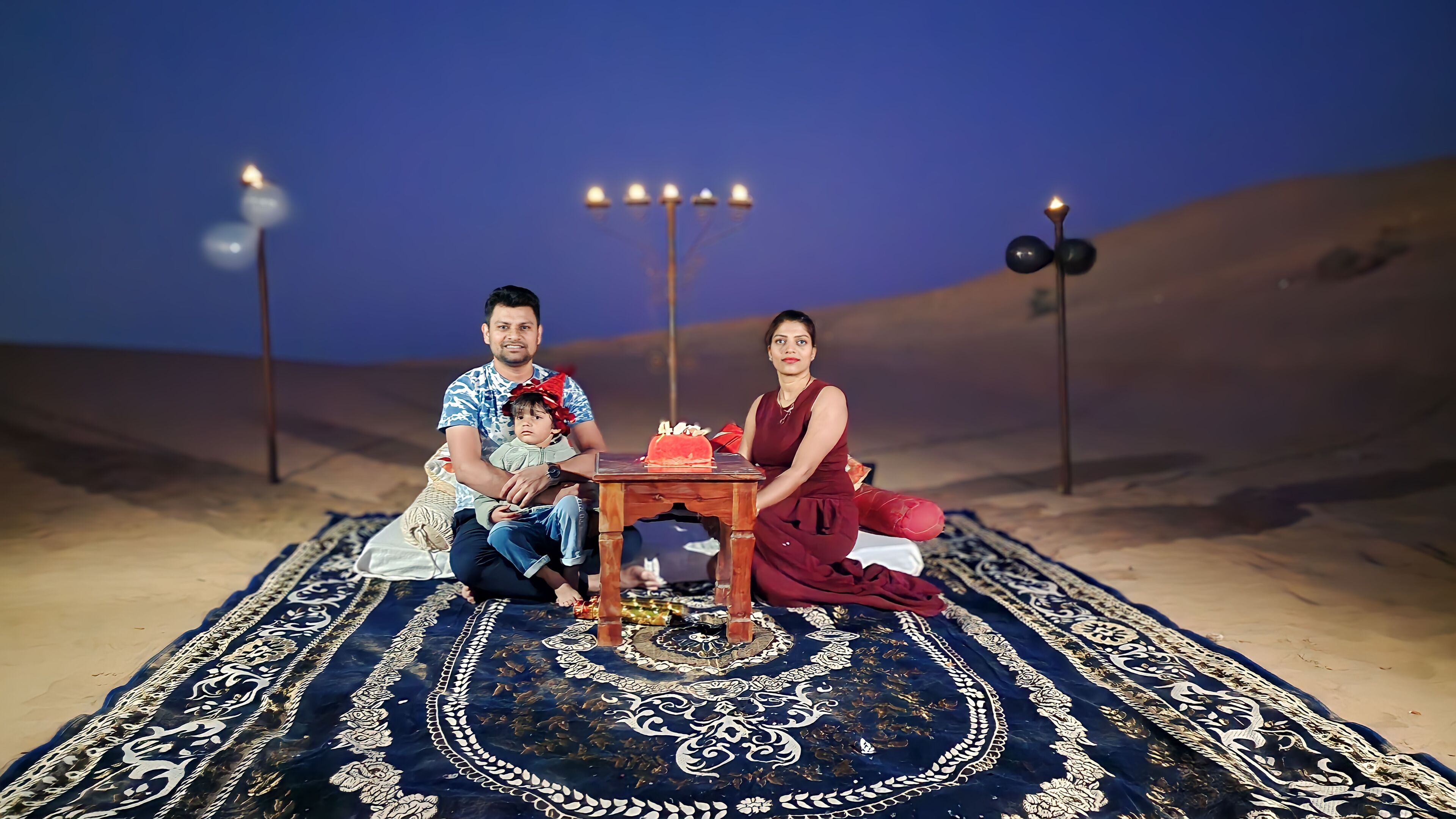 Una familia sentada sobre una alfombra estampada con una mesa en el centro, frente a un paisaje desértico con luces decorativas.