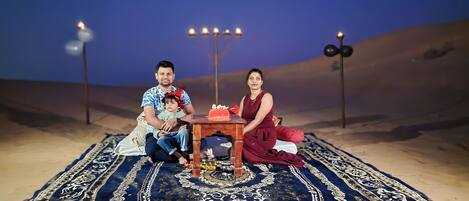 Una familia sentada sobre una alfombra estampada con una mesa en el centro, frente a un paisaje desértico con luces decorativas.