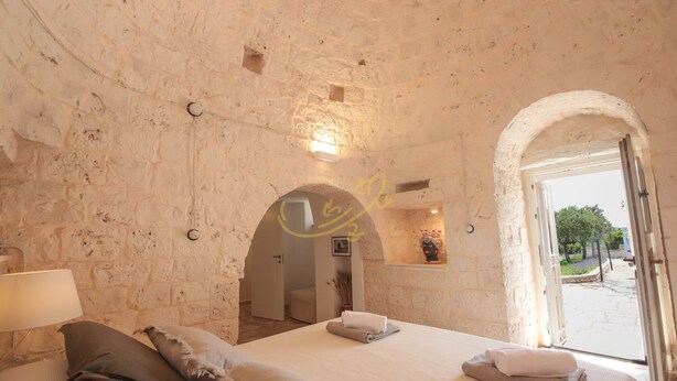 A bedroom with a stone wall, a bed with pillows, a nightstand, and a window with an open door leading outside. - TD Trulli Benedetta w Pool in Peaceful Nature (Castellana Grotte)