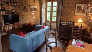 A rustic room with a stone wall, a blue sofa, a wooden TV stand, and a bookshelf filled with books and DVDs. - Natuurhuis (Orliac)