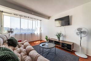 A modern living room with a sofa, a coffee table, a TV, and two standing fans. - Spacious 2BR Apartment in Prime Location with Pool (Las Condes)