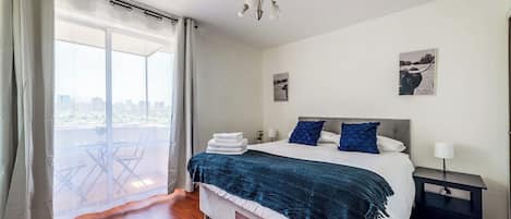 Un dormitorio con cama, mesita de noche, lámpara, ventana con cortinas y vistas al exterior.
