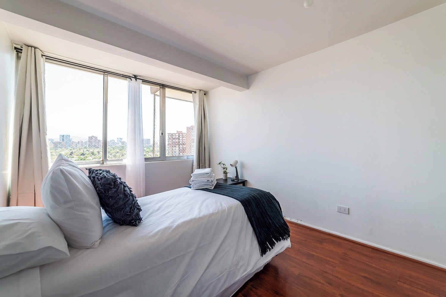 Ein Schlafzimmer mit einem Bett, einem Nachttisch, einer Lampe und Blick auf die Stadt durch große Fenster.