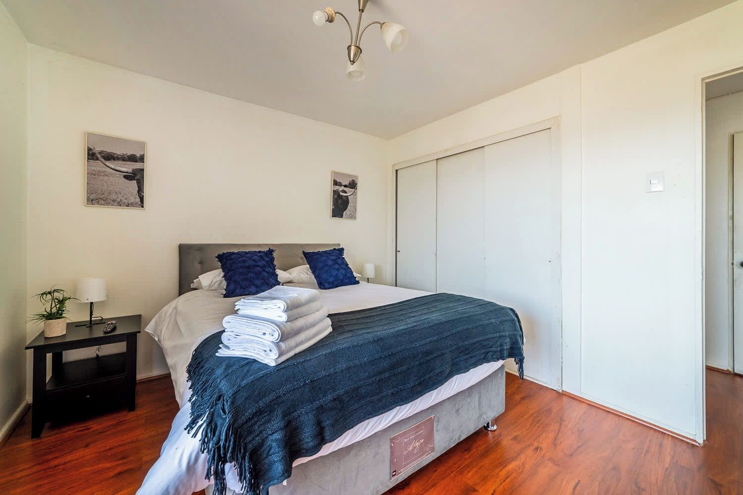 Een slaapkamer met een bed, nachtkastje, lamp en twee aan de muur hangende foto's.