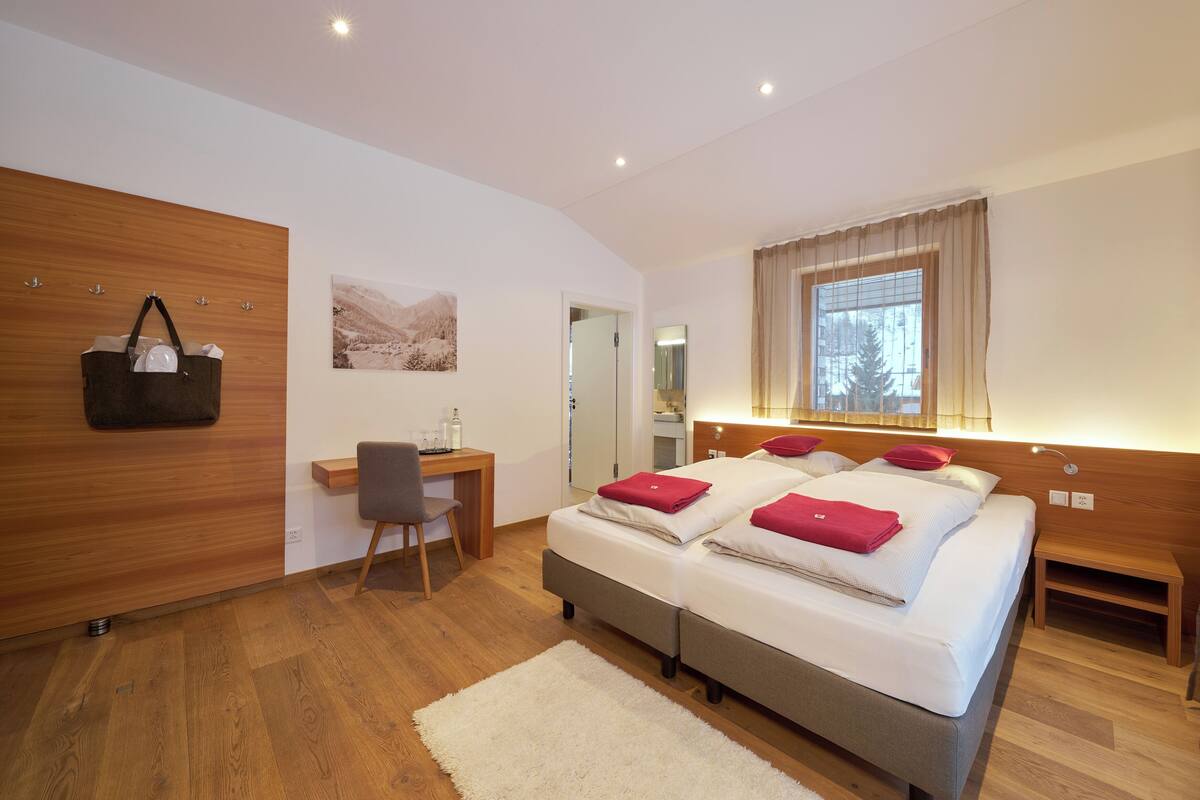 A modern hotel room with a double bed, a desk with a chair, a wooden wardrobe, a large window with curtains, and a wall-mounted bag rack.