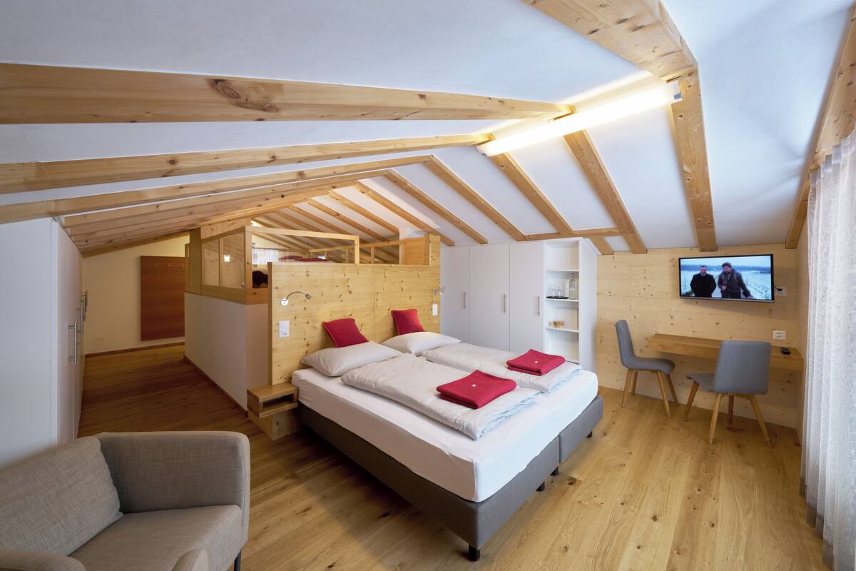 A bedroom with a loft-style ceiling, a bed with red pillows, a desk with a chair, and a TV on the wall.