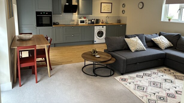 Eine moderne Küche mit grauen Schränken, einem eingebauten Backofen und einer Mikrowelle. Es gibt einen Esstisch mit Stühlen, ein graues Sofa mit Kissen und einen gemusterten Teppich.