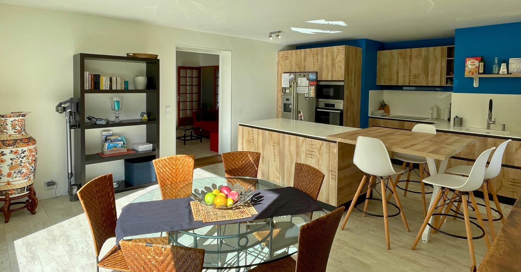 Une cuisine moderne avec des meubles en bois, un îlot central et un coin repas comprenant une table en verre et des chaises en osier.