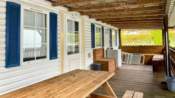 A covered porch with a wooden table and chairs, a small seating area, and a view of a garden. - Spacious mobile home + terrace !, Spacious mobile home + terrace ! (Verchaix)