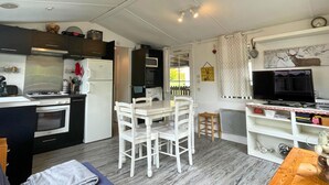 A compact kitchen with a stove, oven, and microwave. There is a dining area with a table and chairs. A television is mounted on the wall. - Spacious mobile home + terrace !, Spacious mobile home + terrace ! (Verchaix)