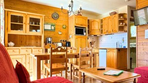 Une cuisine chaleureuse avec des meubles en bois, une table à manger et des chaises. Il y a une horloge au mur et un téléviseur sur le côté droit.