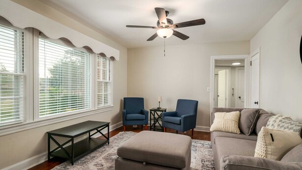 Una sala de estar con un ventilador de techo, dos sillones azules, un sofá gris, un banco y una alfombra estampada.