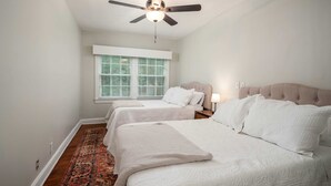 Un dormitorio con dos camas, un ventilador de techo, una ventana con persianas y una alfombra estampada.