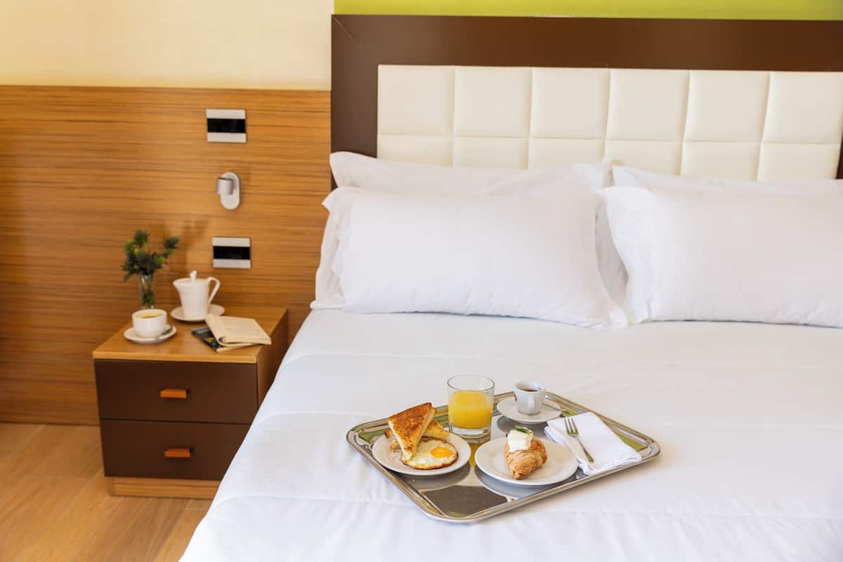 Habitación de hotel con una cama bien hecha, una mesita de noche con una taza de café, y una bandeja de desayuno con tostadas, huevos y jugo.