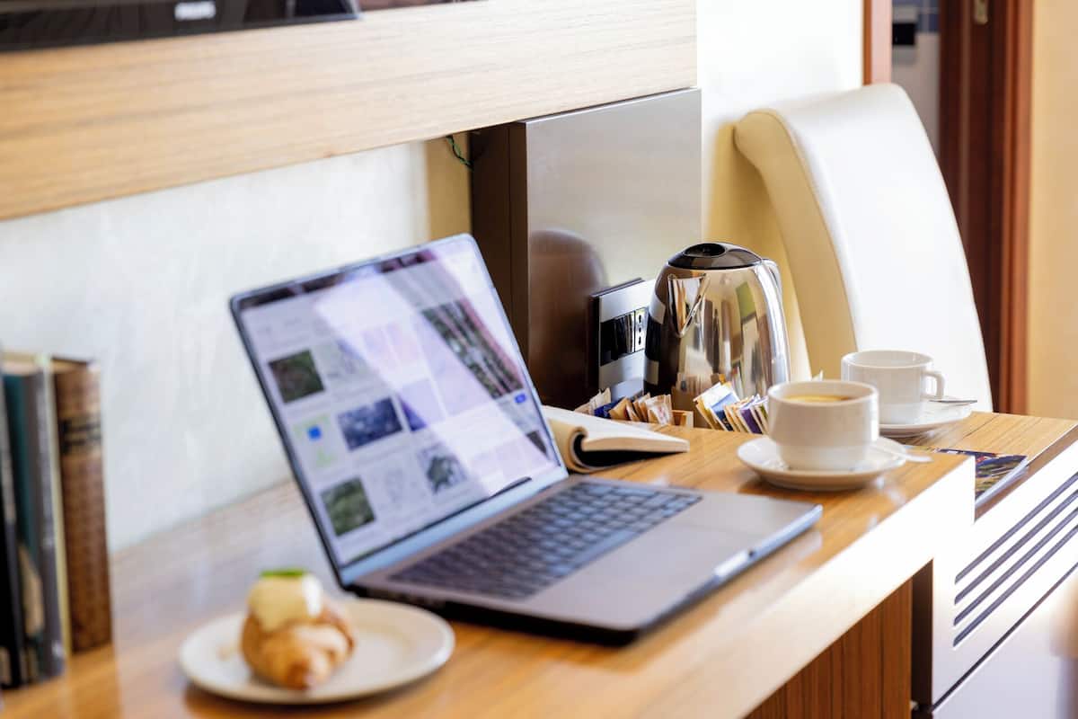 Una laptop sobre un escritorio de madera, con una cafetera, dos tazas y un croissant.