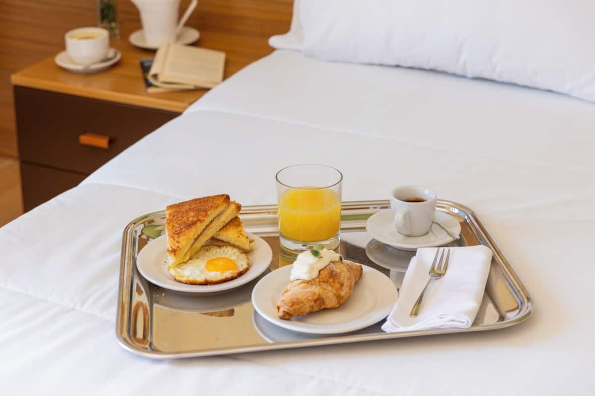 Una cama bien hecha con sábanas blancas, una bandeja con desayuno que incluye tostadas, huevos, jugo y un croissant, y una mesita de noche con un florero y una cafetera.