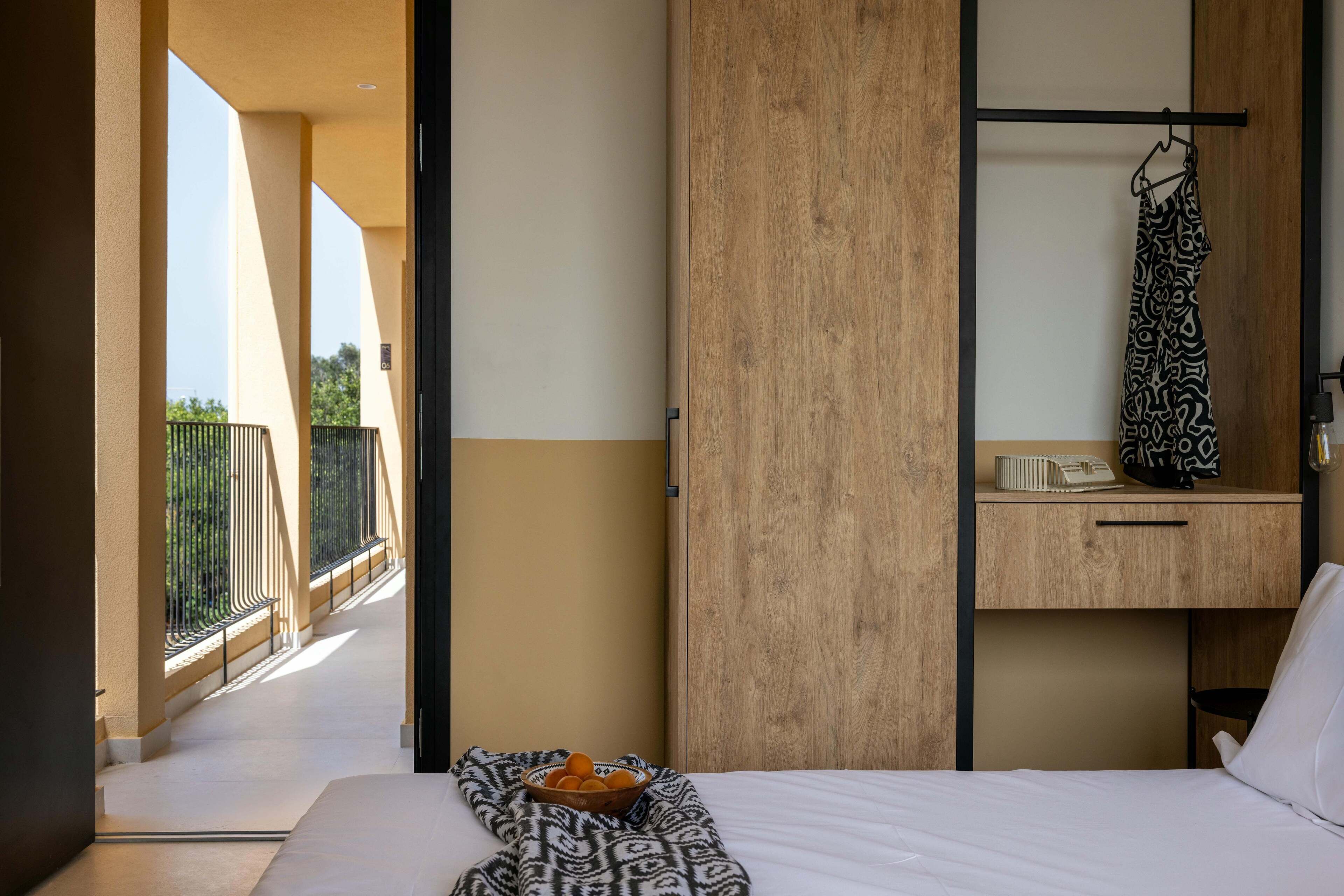 Habitación de hotel con cama, armario de madera, mesita con frutero y vista al balcón exterior.