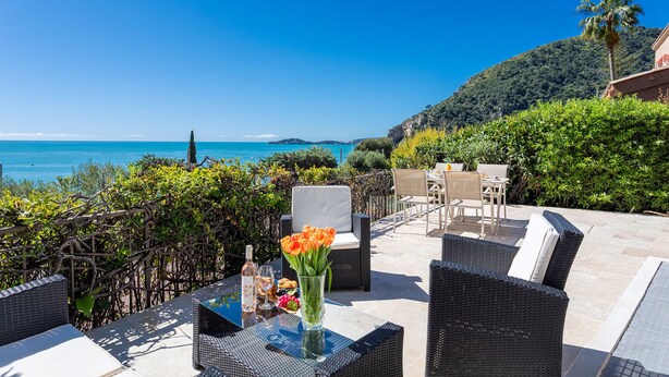 An outdoor patio with wicker furniture, a table with a vase of flowers, and a view of the sea and hills. - VILLA LA PETITE RESERVE VI4416 By Riviera Holiday (Eze)
