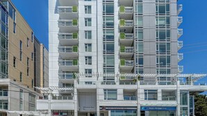 Un immeuble à plusieurs étages doté d’une architecture moderne, avec des balcons agrémentés de jardinières, de grandes fenêtres et une entrée au niveau du sol, ainsi qu’une enseigne indiquant la présence d’un restaurant.
