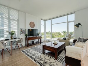 A modern living room with a large TV, a sofa, a coffee table, and a dining area with chairs and a table. - The Bohemian, min 30-Day Stays with Breathtaking Views and Home Office! (Victoria)