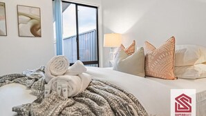 Ein Hotelzimmer mit Bett, Kissen, Decke und Handtüchern. Es gibt ein Fenster mit Vorhängen, einen Nachttisch mit Lampe sowie ein Kunstwerk an der Wand.
