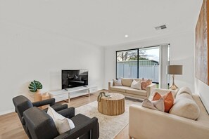 A modern living room with a flat-screen TV, a sofa, a coffee table, and a large window with a view of a fence.