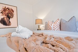 A neatly made bed with a white comforter and a beige blanket, a bedside lamp, patterned pillows, and a framed artwork on the wall.