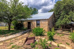Une maison en pierre avec un toit en métal, un jardin avec un chemin en pierre et une brouette en métal.