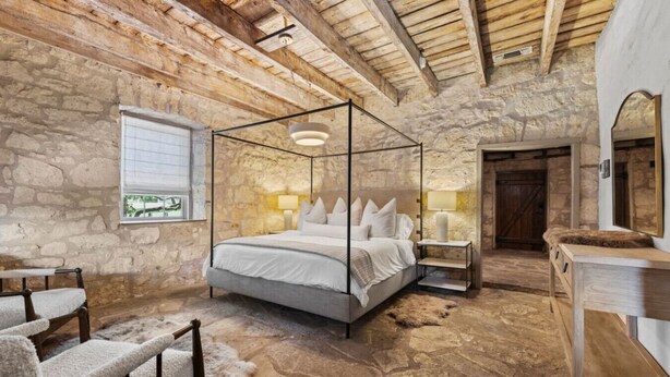 Une chambre aux murs de pierre, dotée d’un lit à baldaquin, d’une commode en bois et d’une fenêtre avec des stores.