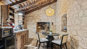 Une cuisine rustique aux murs de pierre, avec des poutres en bois, une table ronde entourée de chaises et un luminaire suspendu.