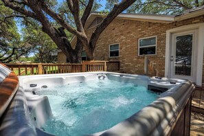 Een hottub in een achtertuin met een huis van baksteen, een houten terras en grote bomen.
