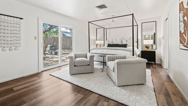 Un dormitorio moderno con cama con dosel, una mesita redonda, dos sillones, un espejo grande y vistas al exterior a través de una puerta corrediza de cristal.