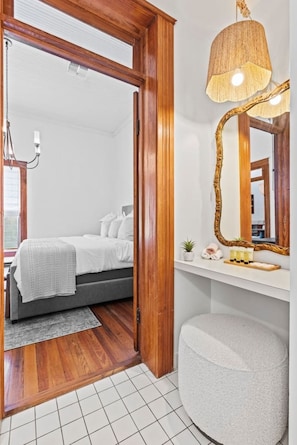 Un dormitorio moderno con puerta de madera, cama con ropa de cama blanca, lámpara de noche, espejo y una pequeña planta en la mesita.