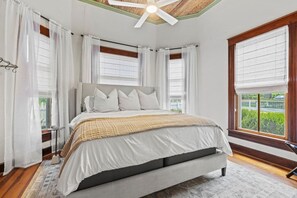 A bedroom with a bed, white curtains, a ceiling fan, and large windows.