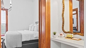 A modern bedroom with a wooden door, a bed with white bedding, a bedside lamp, a mirror, and a small plant on the nightstand. - Danz Haus | Historic Home | Baron's Creek (Fredericksburg)