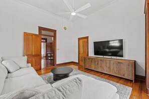 Una sala de estar con un sofá blanco, un mueble de televisión de madera, un ventilador de techo y una puerta de madera.
