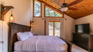 A bedroom with a large bed, wooden headboard, a ceiling fan, a television on a dresser, and large windows with curtains. - Grandview Getaway (Deep Gap)