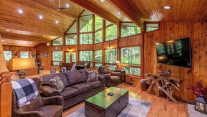 A spacious cabin interior with a large wooden ceiling, multiple windows offering views of the forest, a leather sofa, and a flat-screen TV mounted on the wall. - Gait to Paradise (Boone)