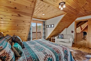 A cozy attic bedroom with a sloped ceiling, a large bed with patterned bedding, a chair, and a view of the outdoors. - Ponderosa 1887 (Vilas)