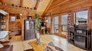 Ein rustikales Blockhaus im Inneren, mit Holzwänden und -decken, einem Essbereich mit Tisch und Stühlen, einer Küche mit Kühlschrank und einem großen Fenster mit Jalousien.
