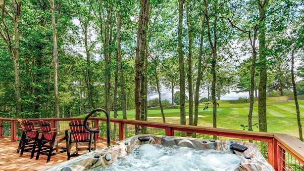 A hot tub on a wooden deck with chairs, surrounded by a forest and a golf course in the background. - Love to Beech (Banner Elk)