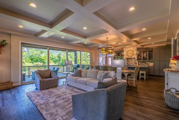 A spacious living area with a large sofa, armchairs, and a dining table. There is a balcony with a view of greenery. - Natures Blessings at Elk Creek (Banner Elk)