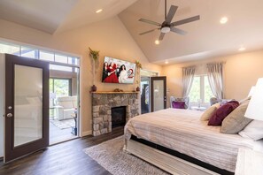 A bedroom with a stone fireplace, a ceiling fan, a bed with multiple pillows, and a large window. - Natures Blessings at Elk Creek (Banner Elk)