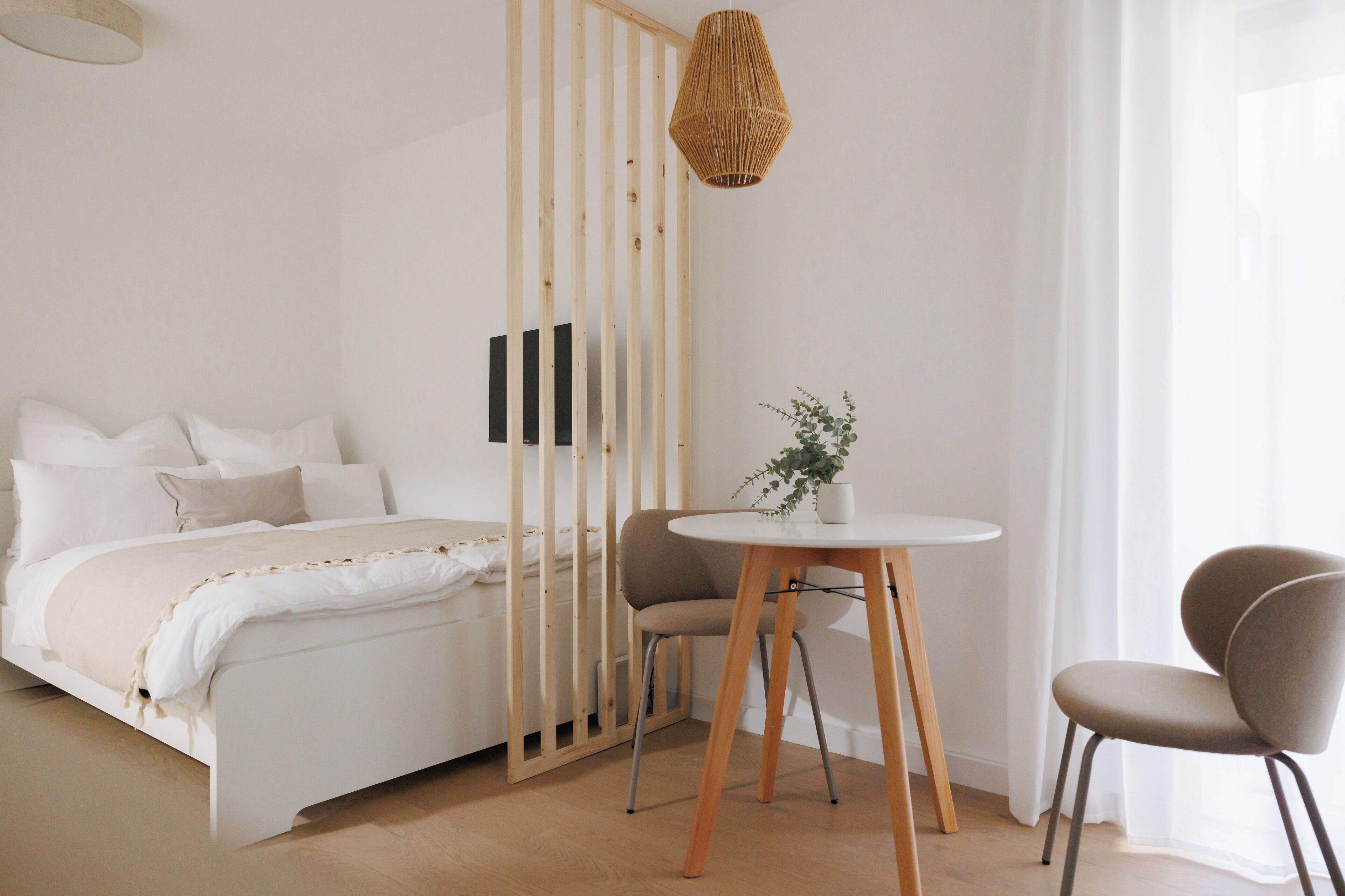 A bedroom with a bed, a chair, a small table, a hanging light, and a window with curtains.
