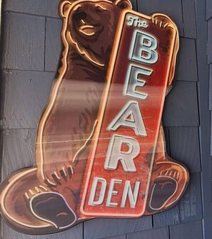 A bear-shaped neon sign with the text "The Bear Den" on a wooden wall.