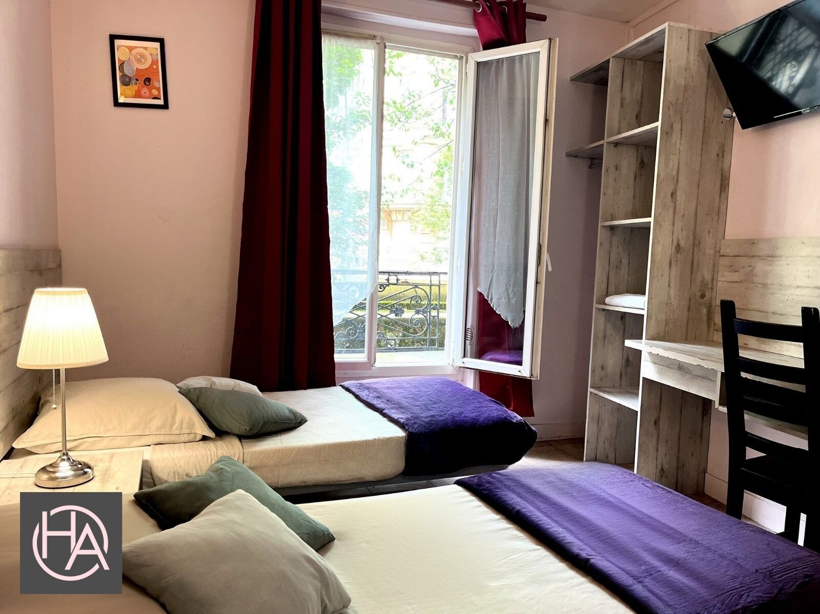 Un dormitorio con dos camas, una mesita de noche con lámpara, una ventana con cortinas y un televisor fijado a la pared.