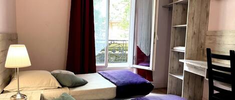 A bedroom with two beds, a bedside table with a lamp, a window with curtains, and a TV mounted on the wall.