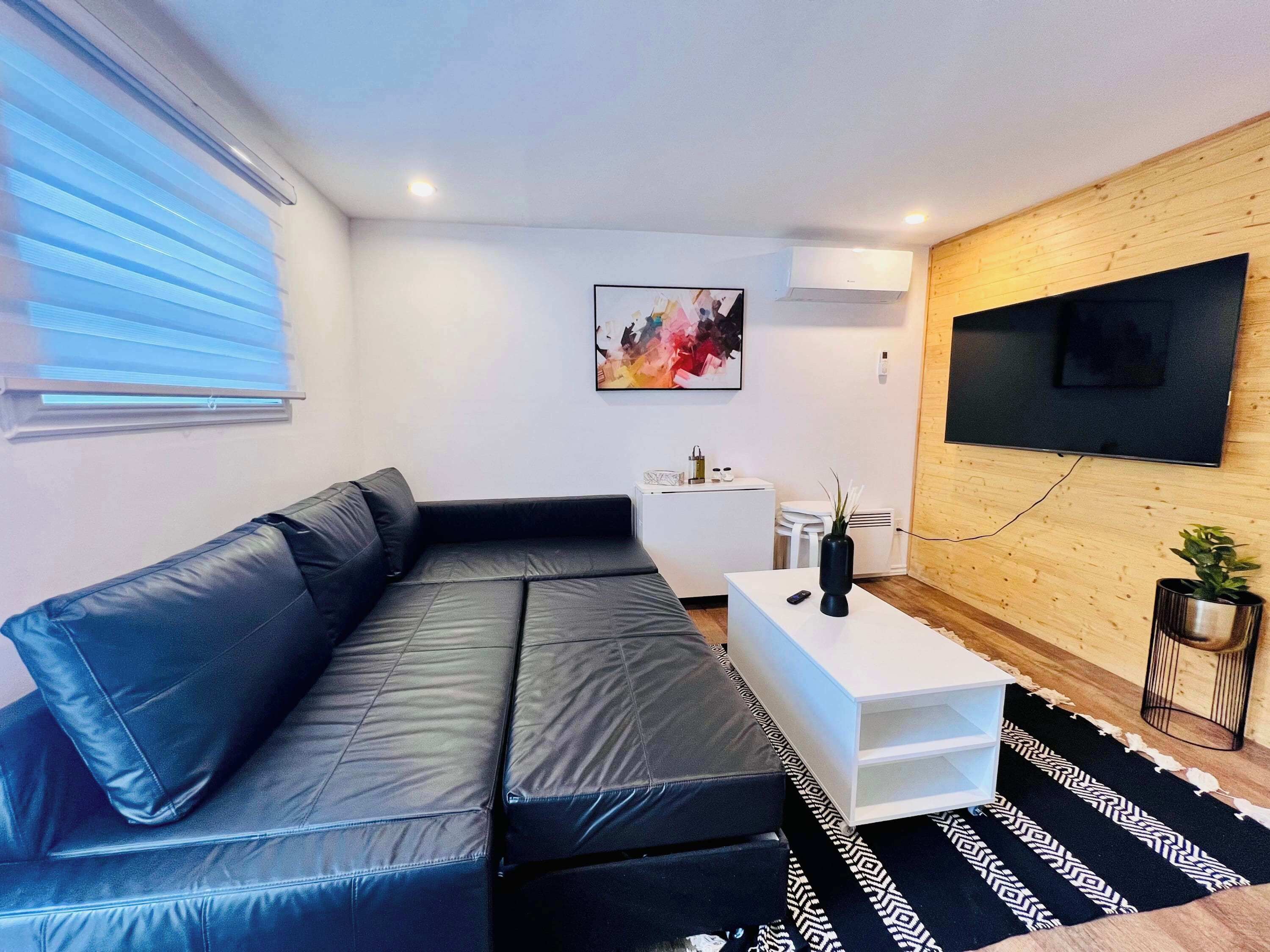 Un salon moderne avec un canapé en cuir noir, une table basse blanche, un téléviseur fixé au mur et un petit meuble blanc.